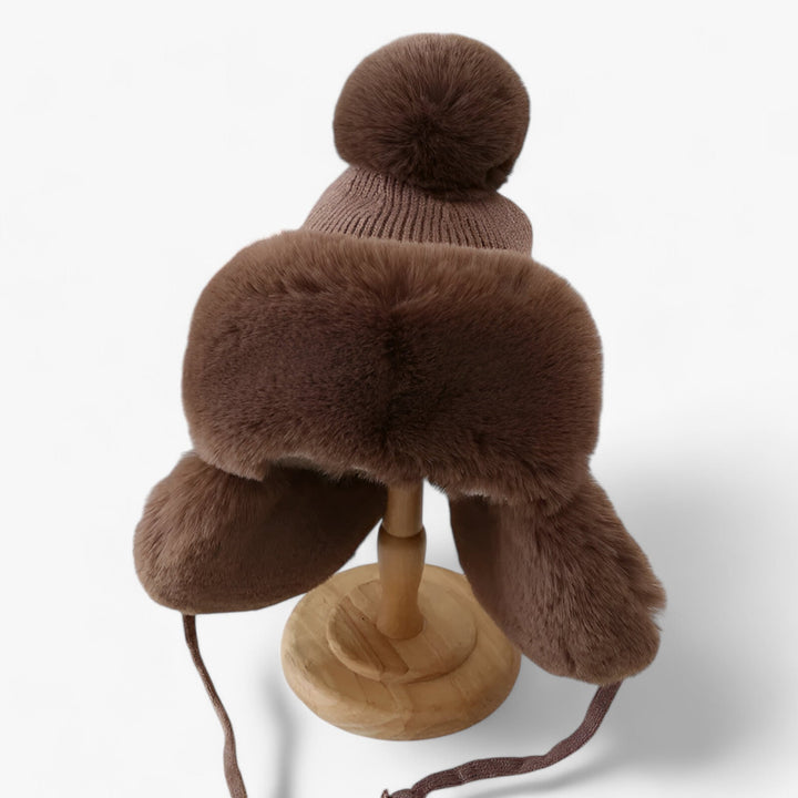 Braune Wintermütze mit Kunstfell, Bommel und Ohrenklappen auf Holzständer.