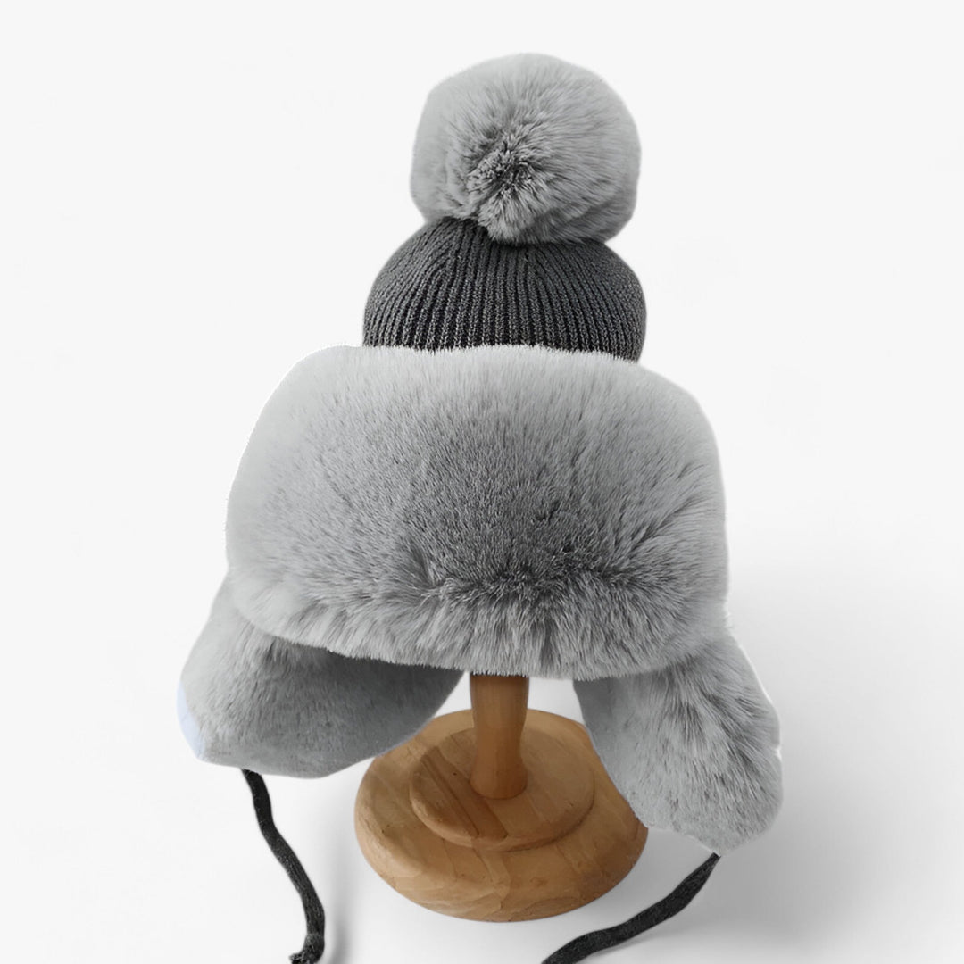 Graue Wintermütze mit Kunstfell, Bommel und Ohrenklappen auf Holzständer.