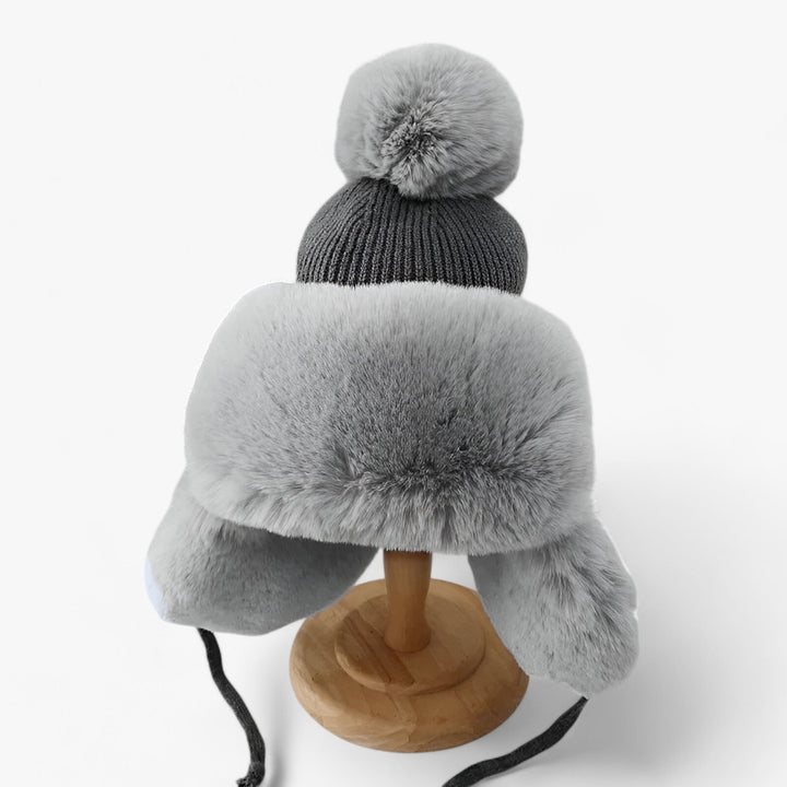 Graue Wintermütze mit Kunstfell, Bommel und Ohrenklappen auf Holzständer.