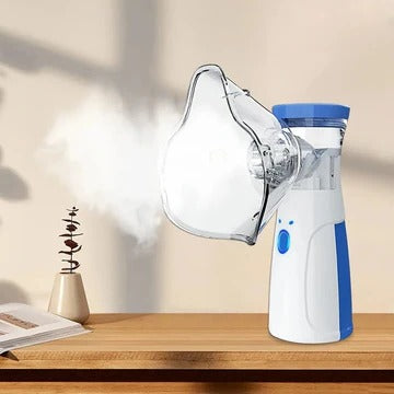 Bílomodrý ruční nebulizér s nasazenou průhlednou maskou chrlí mlhu na dřevěném stole vedle otevřené knihy a malé rostliny. 1