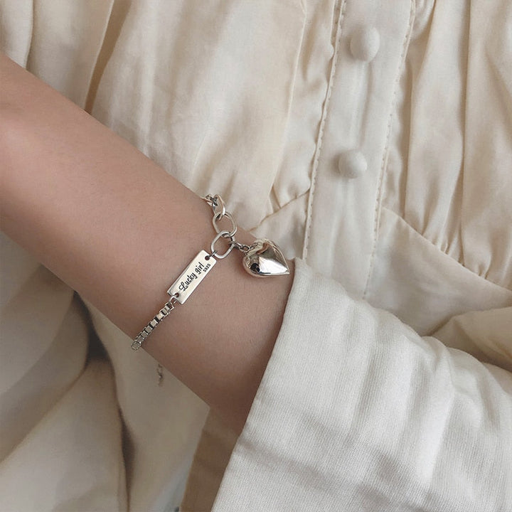 Silberarmband mit Herzanhänger und Gravur auf beigem Hemdärmel.