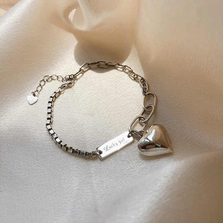 Silberarmband mit Herzanhänger, graviert mit "Lucky girl", auf beigem Stoffhintergrund.
