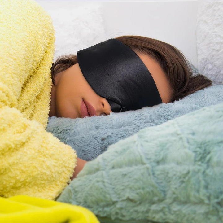 Frau mit schwarzer Schlafmaske, gelbem Bademantel, schläft auf weichem Kissen.