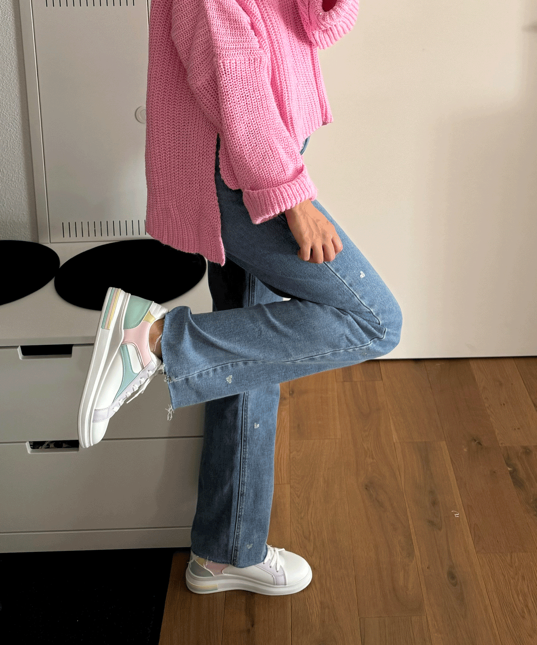 Frau in rosa Pullover und blauen Jeans trägt weiße Sneaker mit pastellfarbenen Details.