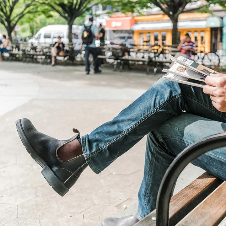Mann in Jeans und schwarzen Stiefeln liest Zeitung auf Parkbank.