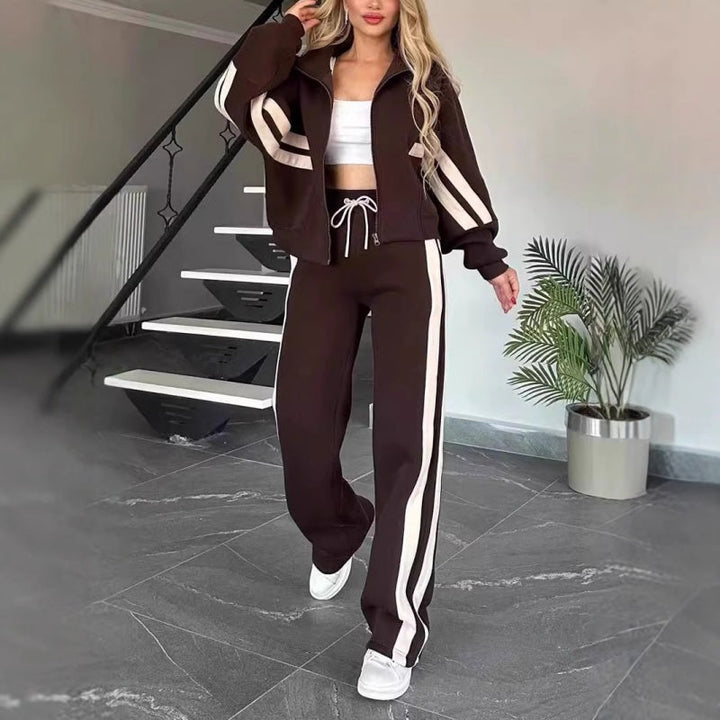 Souprava v tmavě hnědé barvě s krémovými pruhy, modelka kráčí po místnosti, bunda na zip a široké kalhoty s bílou šňůrkou. 8