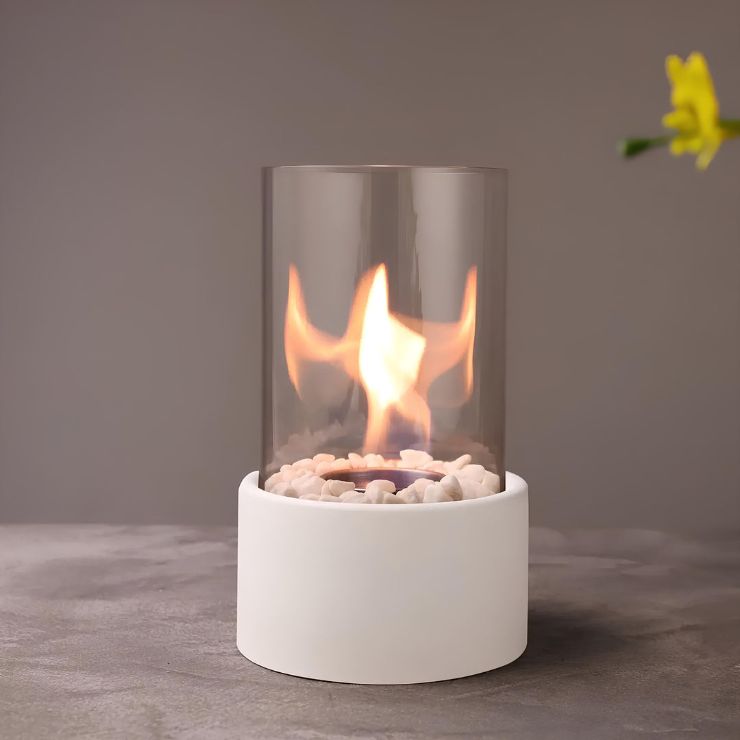 Moderner Tischkamin mit Glaszylinder, weißem Sockel und dekorativen Steinen, ideal für Innenräume.