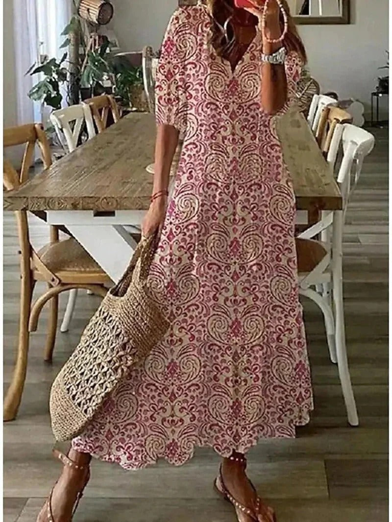 Frau in rosa Maxikleid mit Paisley-Muster, Strohtasche, Sandalen, Esszimmer im Hintergrund.
