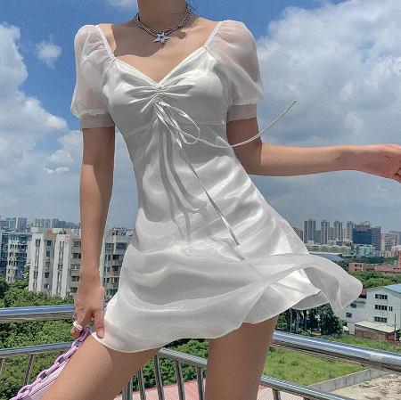 Frau in weißem Sommerkleid mit Puffärmeln auf Balkon vor Stadtlandschaft.