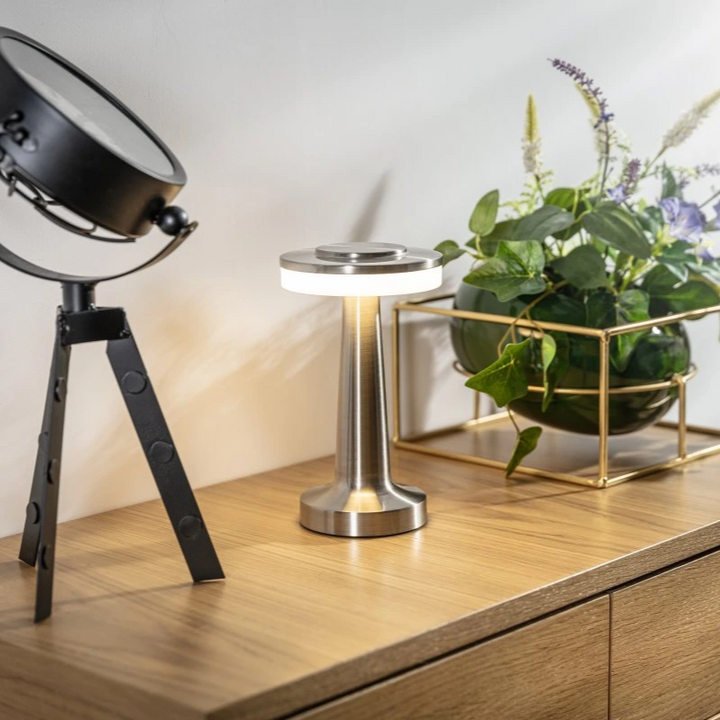 Moderne Tischlampe aus Metall auf Holzoberfläche neben Pflanze in goldener Halterung.