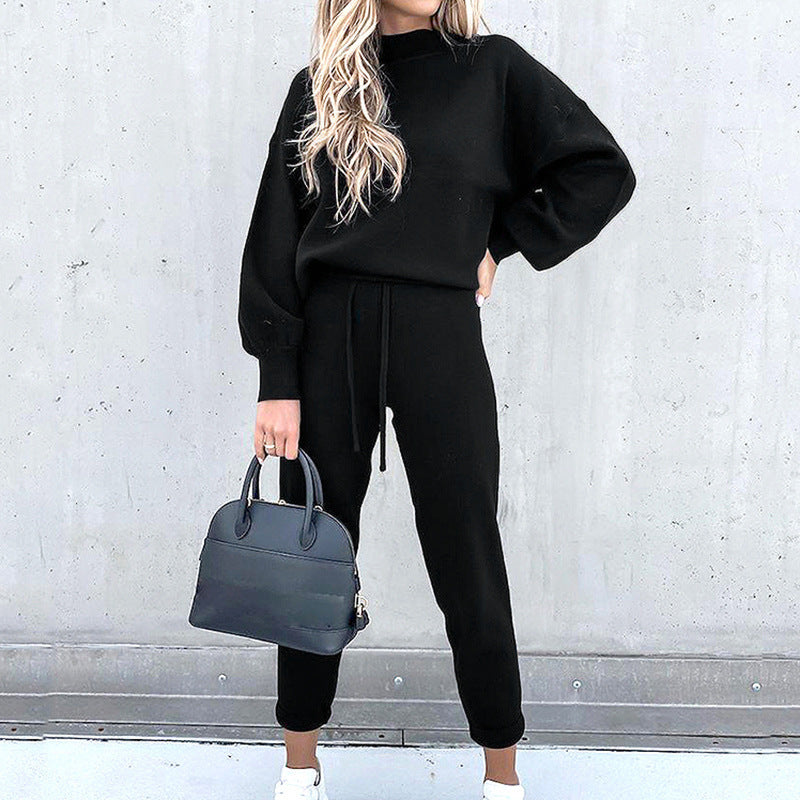 Frau in schwarzem Hausanzug, zweiteilig, mit Rollkragen und High-Waist-Hose, hält Handtasche.