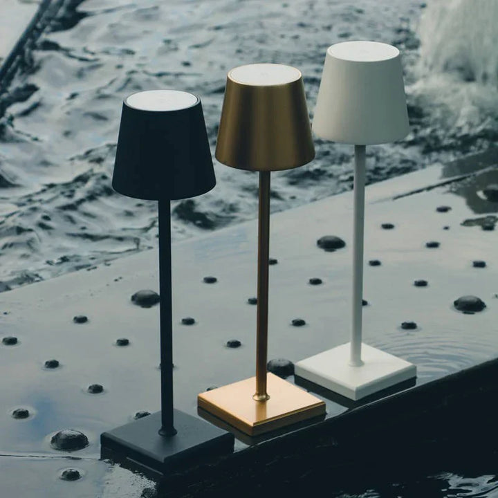 Drei Poldina Pro Tischlampen in Schwarz, Gold und Weiß auf Metalloberfläche am Wasser.